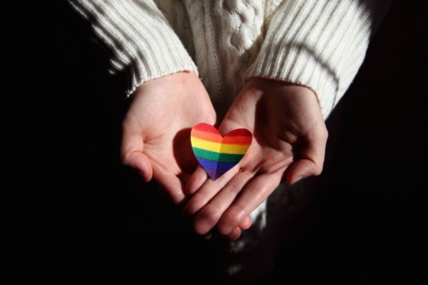 Une personne tient dans ses mains un coeur en papier aux couleurs de l'arc-en-ciel en soutien aux personnes LGBTQIA+.